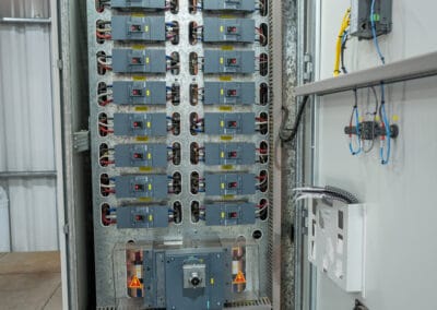 Interior de quadro elétrico industrial aberto com colunas de disjuntores modulares, dispositivo principal de manobra com alertas de alta tensão, fiacao encaminhada em calhas e iluminação interna por LED.